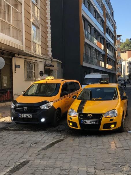 Aydın 7 kişilik taksi: Arnavut kaldırımlı Aydın sokağında Dacia Lodgy geniş araç ve Volkswagen Golf sarı taksiler